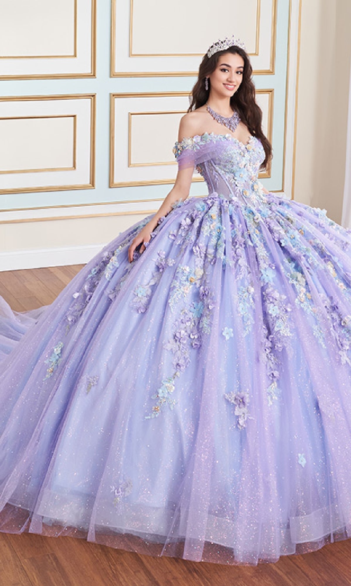 A woman wearing the Quinceanera Dress PR30175 By Princesa—a sparkling lavender gown with beading and 3D floral accents—stands crowned with a tiara in a decorated room with light walls and wooden flooring.